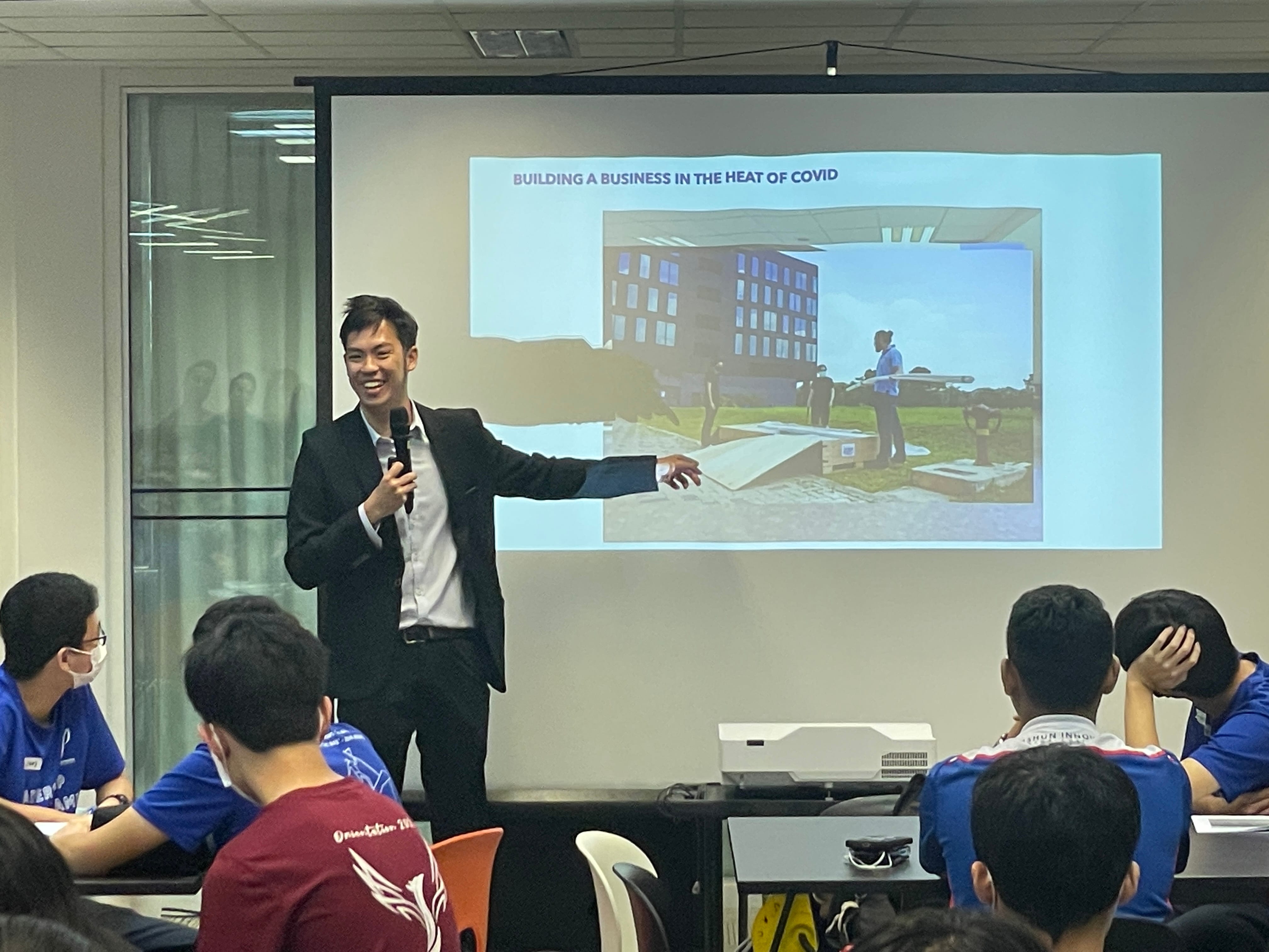 A man in a black suit holds a microphone and points to a projected slide titled "Building a Business in the Heat of Covid."